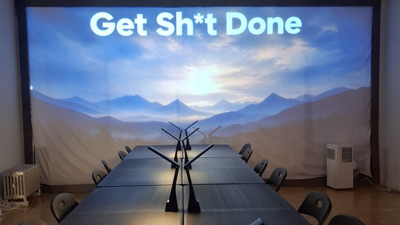 The Get Sh*t Done Club room: long black tables and chairs facing a mountain backdrop, with Get Sh*t Done projected across the top.